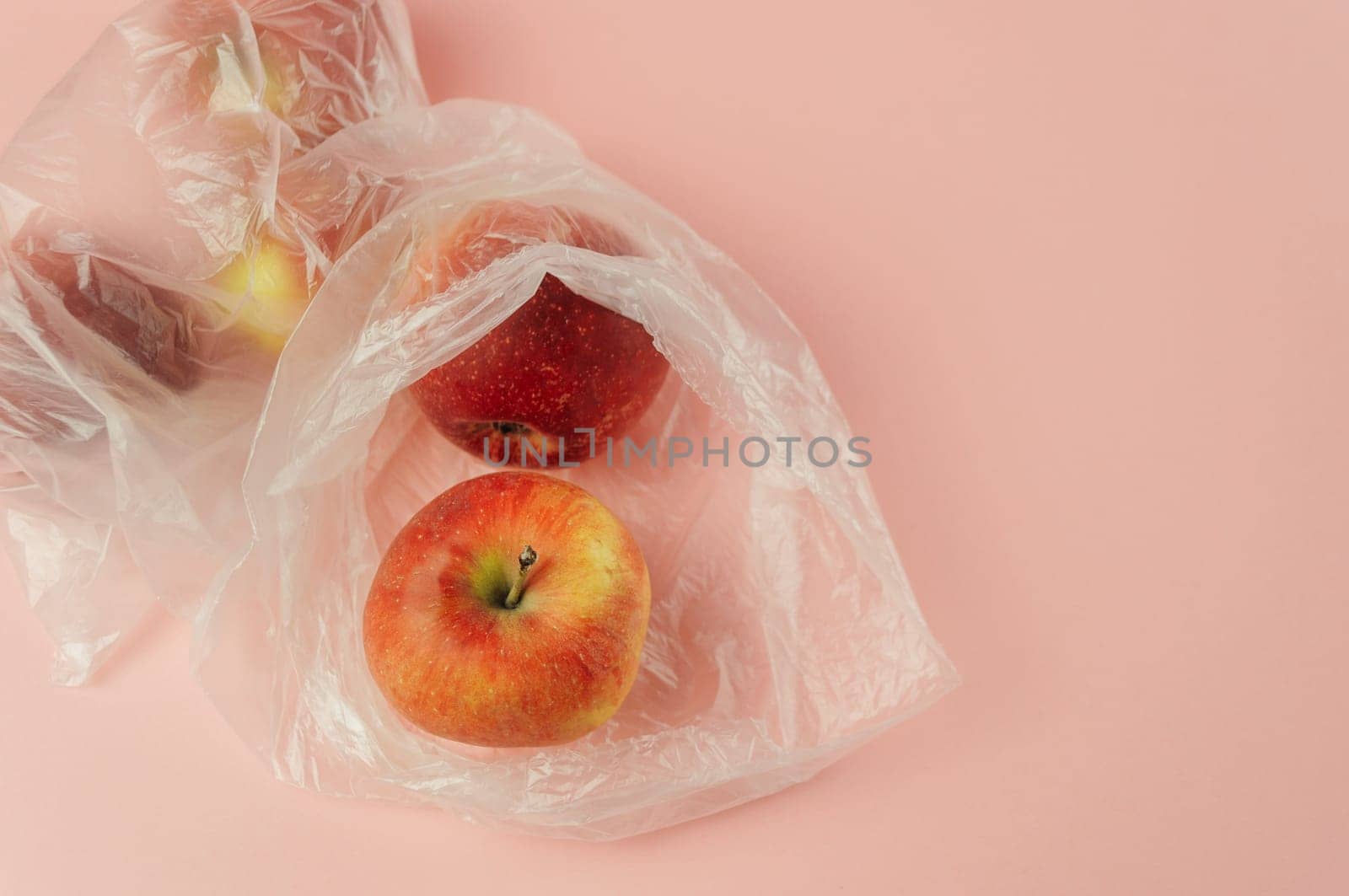 A plastic bag with a red apple inside. The bag is placed on a pink background. Concept of wastefulness and environmental concern, as the plastic bag is used to hold the apple, which is a natural