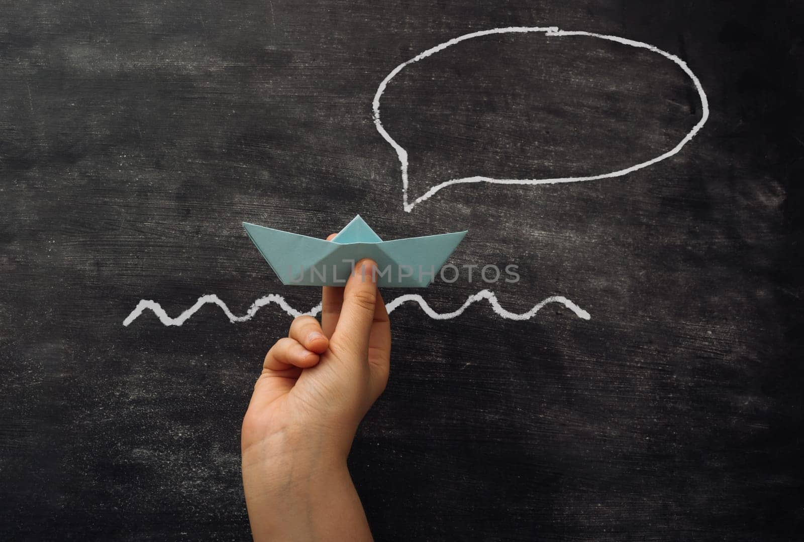 A hand holding a paper boat in front of a chalkboard with a speech bubble. The speech bubble says I am a boat.
