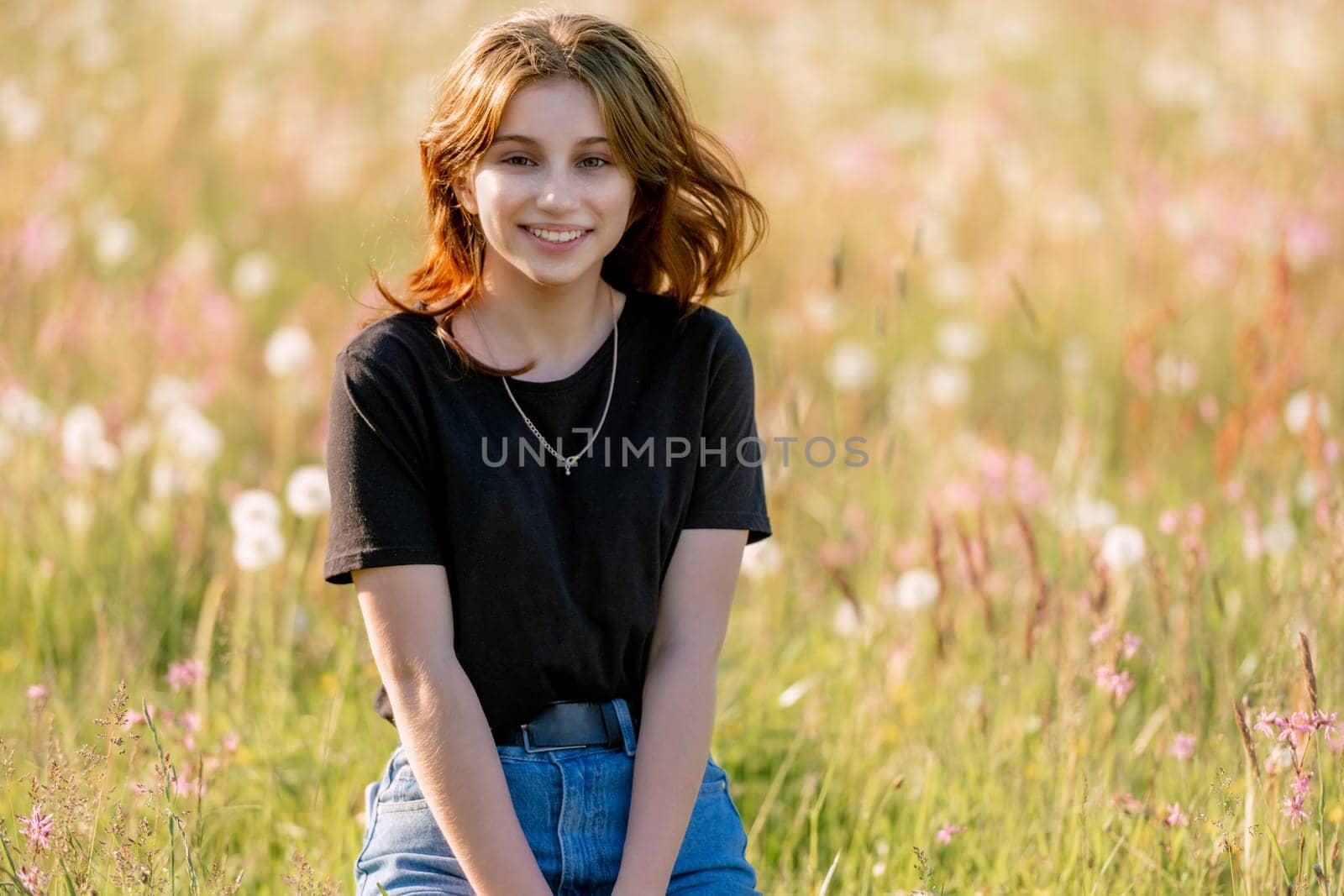 Pretty girl teenager sitting in field with dandelions and smiling outdoors. Beautiful young female person at nature in sunny day portrait