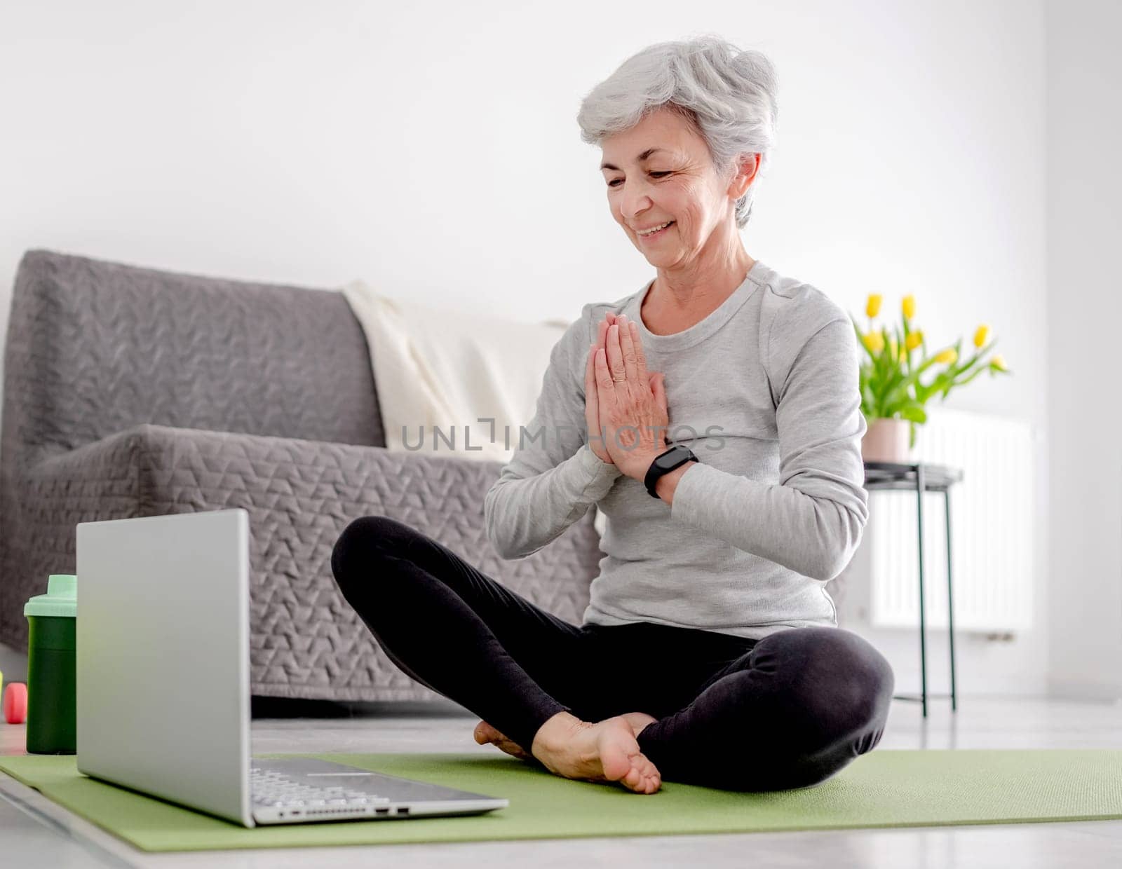 Pleasant 65-Year-Old Woman Greets Her Trainer Through Laptop For An Online Sport Session At Home On A Yoga Mat