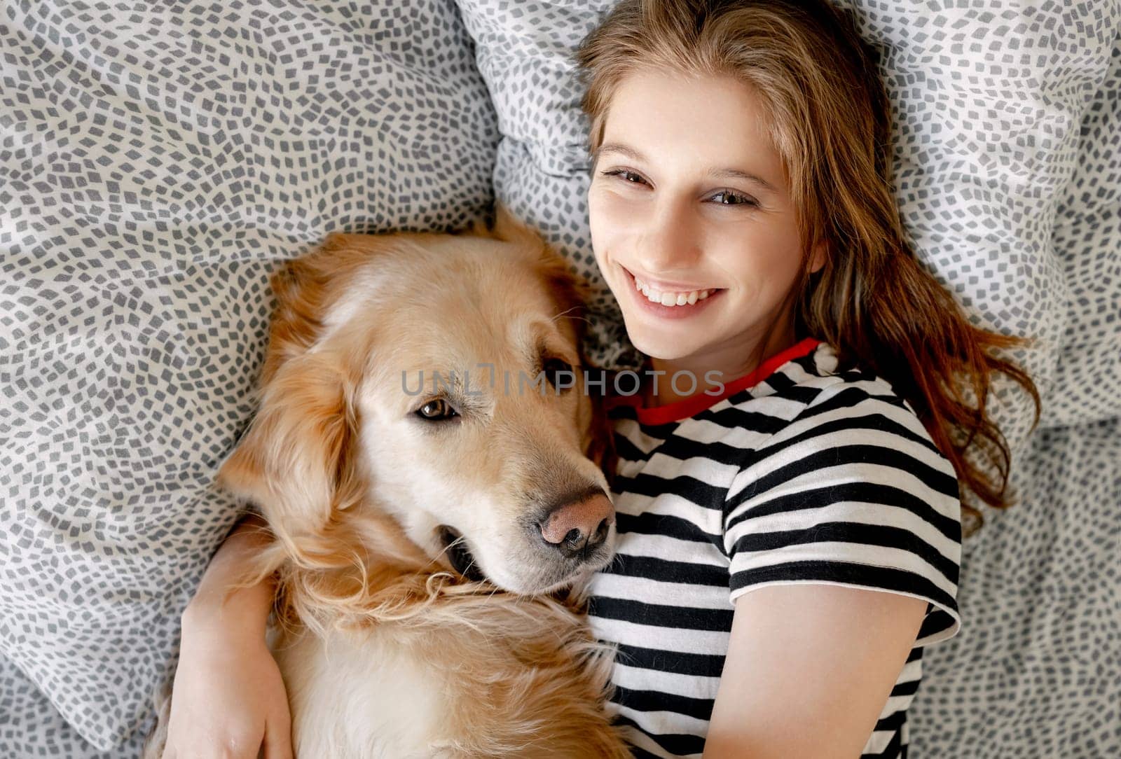 Pretty girl hugging golden retriever dog and smiling lying in bed. Happy teenager with purebred doggy pet labrador resting together