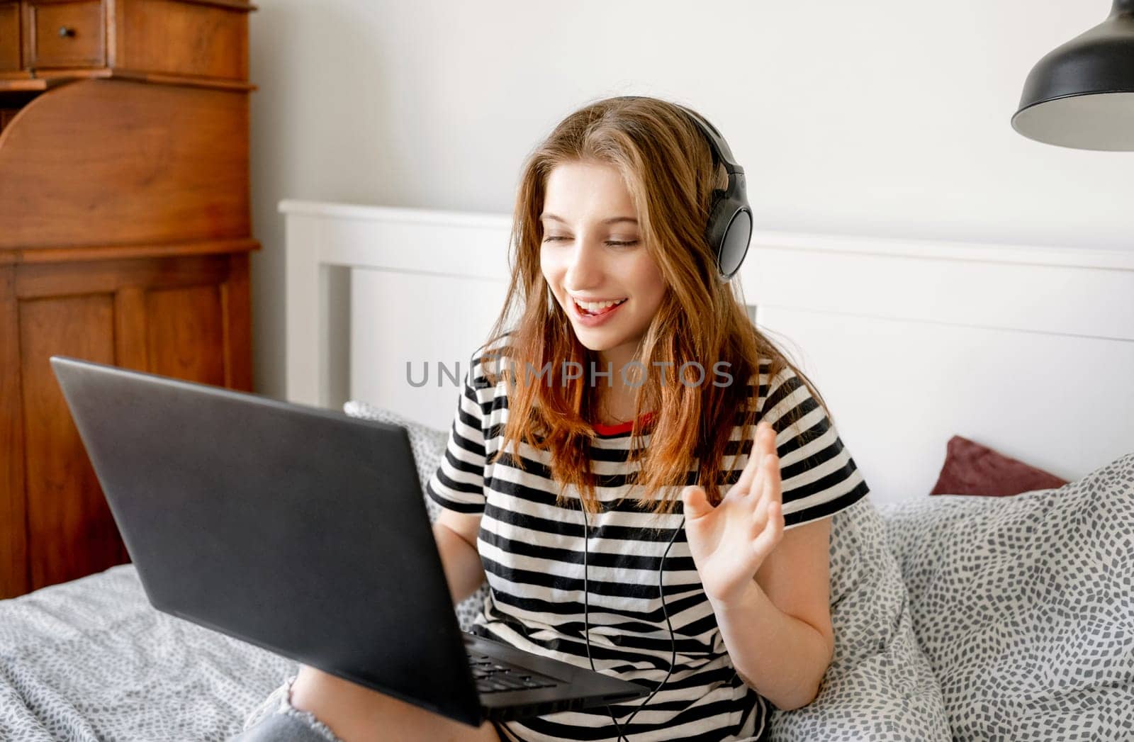 Pretty girl with headphones and laptop in bed during online learning webinar. Beautiful teenager making video call with computer at bedroom