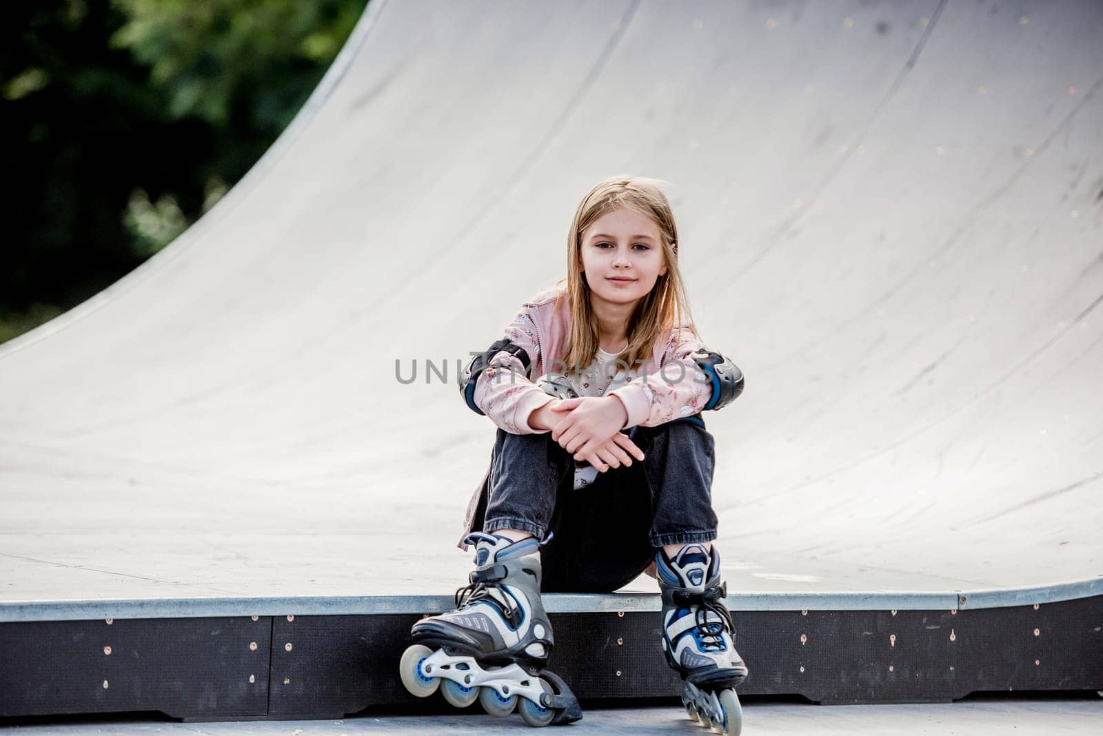 Cute girl roller skater sitting in city park and looking at camera Pretty female preteen kid child posing during rollerskating