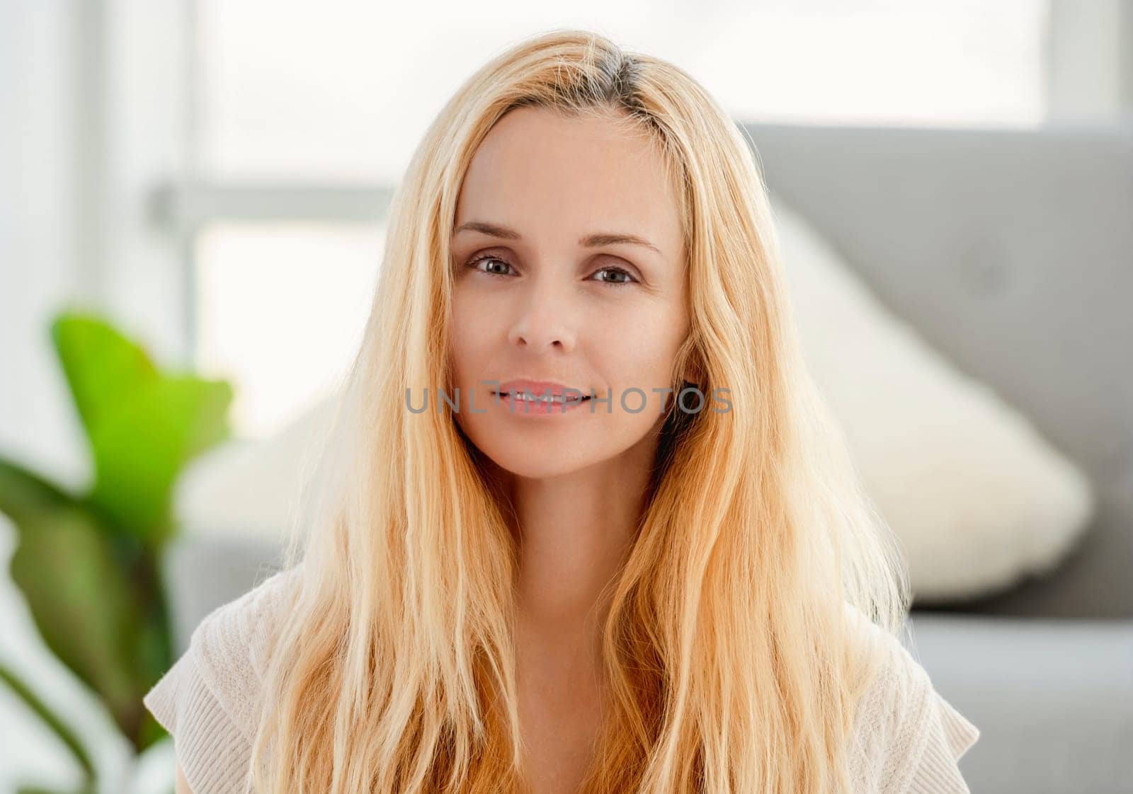 Beautiful young blond girl smiling and looking at the camera at home. Portrait of pretty woman with light blurred interior on background