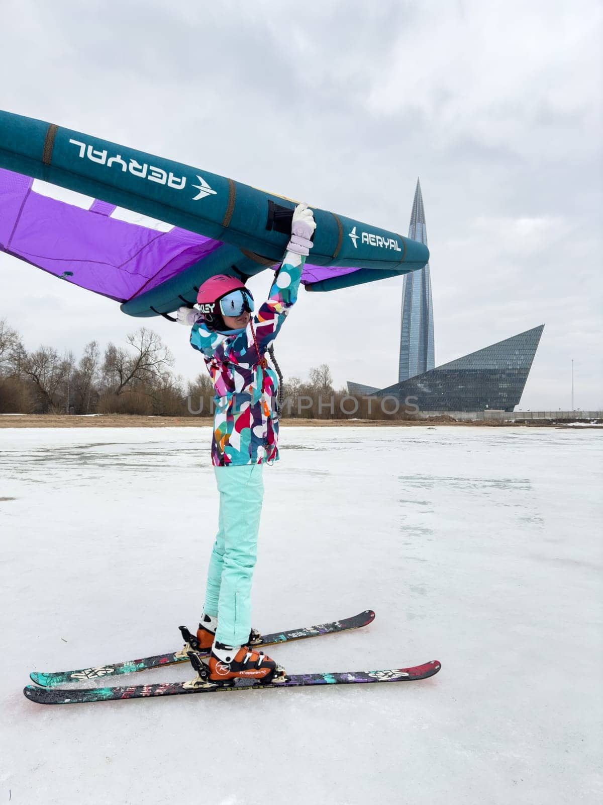 Russia, St.Petersburg, 14 March 2024: A girl in bright colored ski gear is ready to ski on ice with an air wing at cloudy weather, the Lakhta Center skyscraper in the background, helmet and glasses by vladimirdrozdin