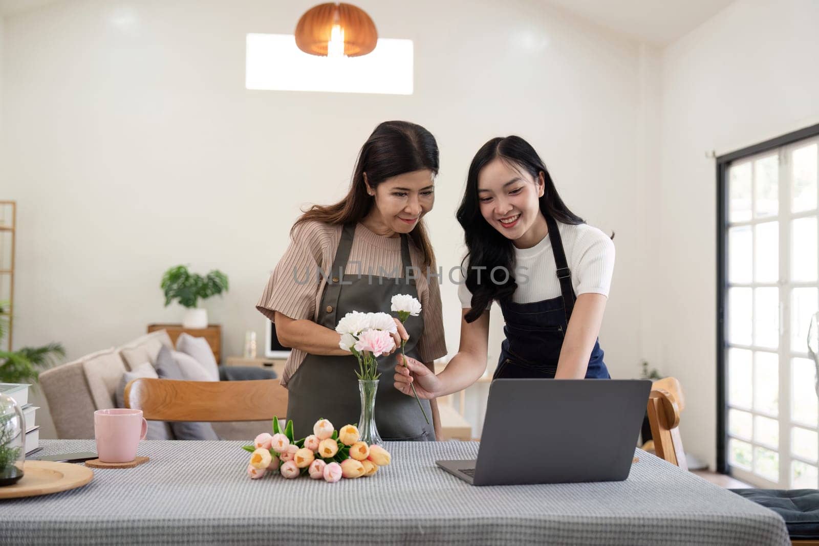 Senior mother and adult daughter happy on the table while arrange flowers in a vase together using laptop computer. lifestyle concept. Happy time together by nateemee