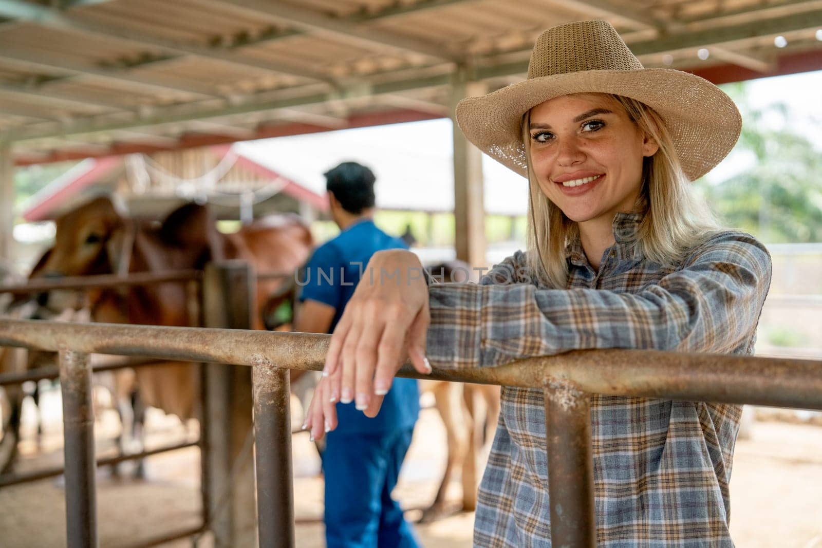 Beautiful caucasian farmer with hat relax near stable railing also look at camera with smiling and the veterinarian also check health of cows in background.