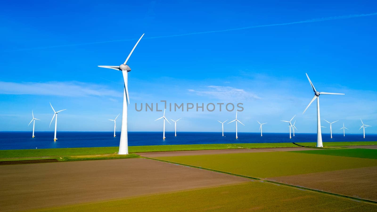 A cluster of windmills stands tall in a field next to the ocean, their blades spinning gracefully in the spring breeze. windmill turbines, green energy, earth day