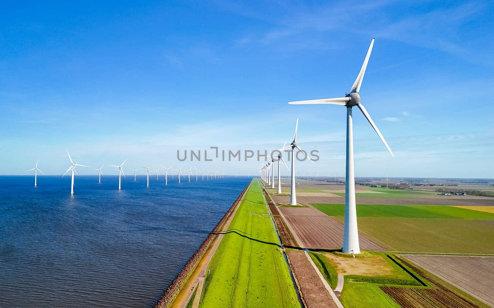 row of wind turbines elegantly spin next to the tranquil body of water in the light of spring by fokkebok