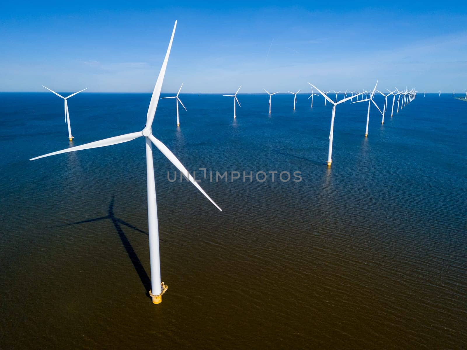 A group of windmills floats gracefully in the Netherlands Flevoland region during the vibrant season of Spring. drone aerial view of windmill turbines green energy in the blue ocean