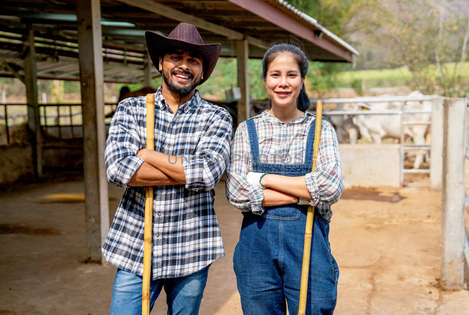 Portrait of Asian man and woman farmers carry broom and stand with arm crossed also look at camera with smiling in front of stable of cow look relax after clean the area. by nrradmin