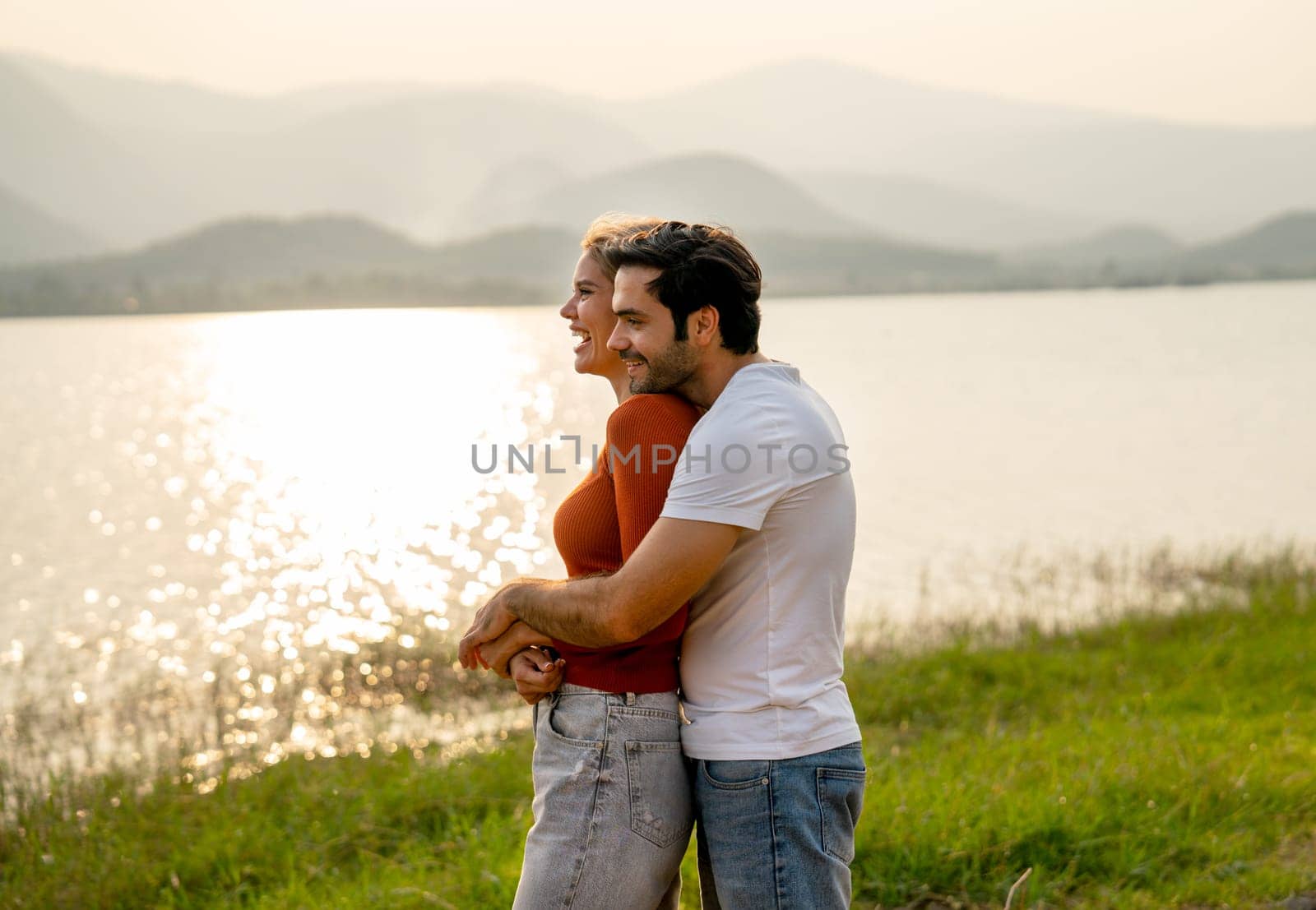 Caucasian man hug woman from the back and stay together near lake with sunset and romance in area of public park. by nrradmin