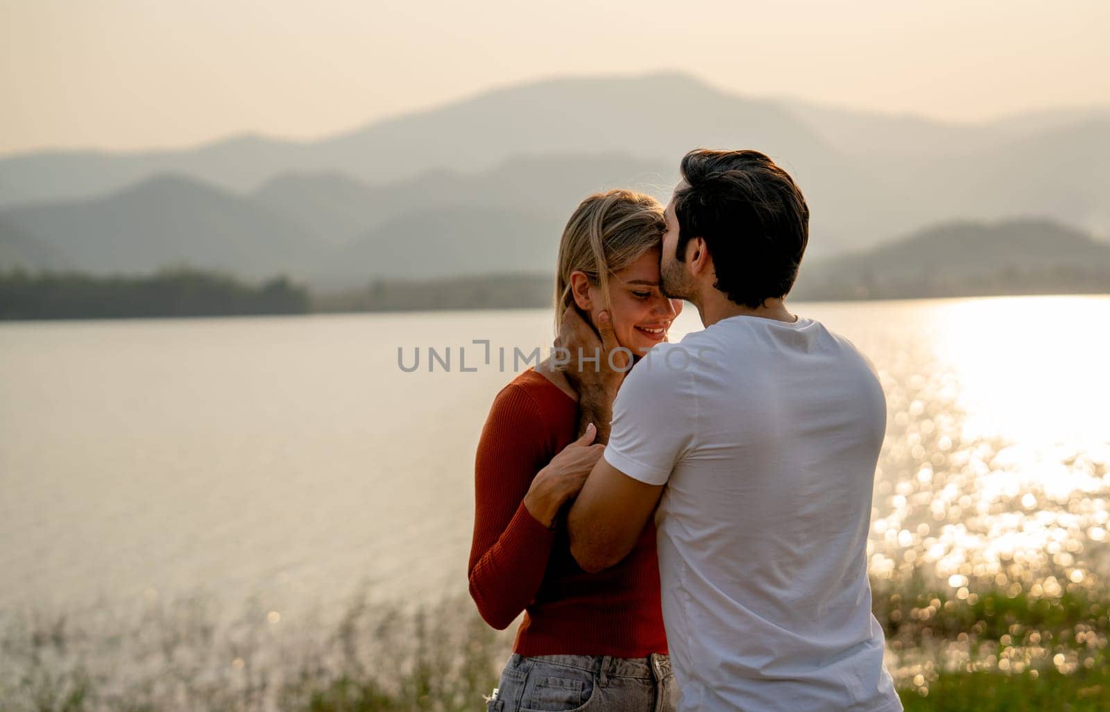 Back of Caucasian man kiss on forehead of beautiful woman with sunset light near lake and look romantic for couple love stay together.