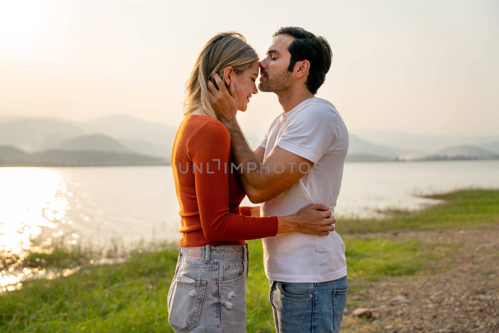 Caucasian man kiss on forehead of beautiful woman with sunset light near lake and look romantic for couple love stay together. by nrradmin