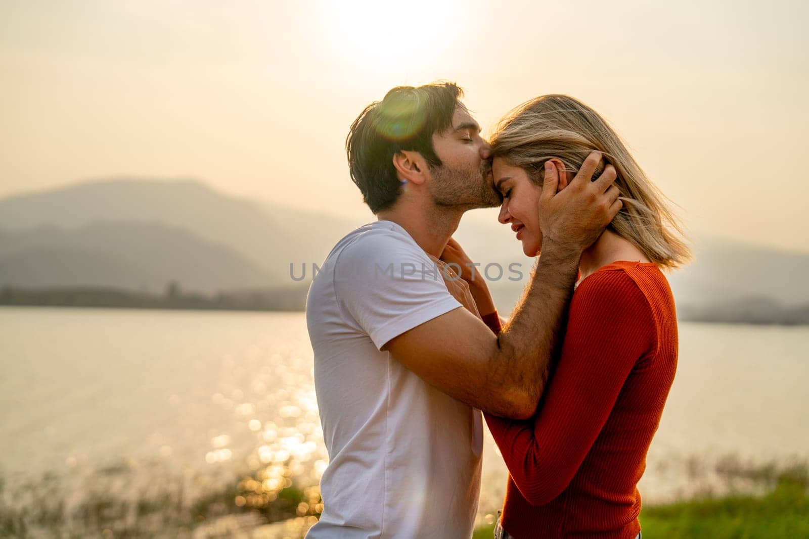 Close up Caucasian man kiss on forehead of beautiful woman with sunset light near lake and look romantic for couple love stay together. by nrradmin