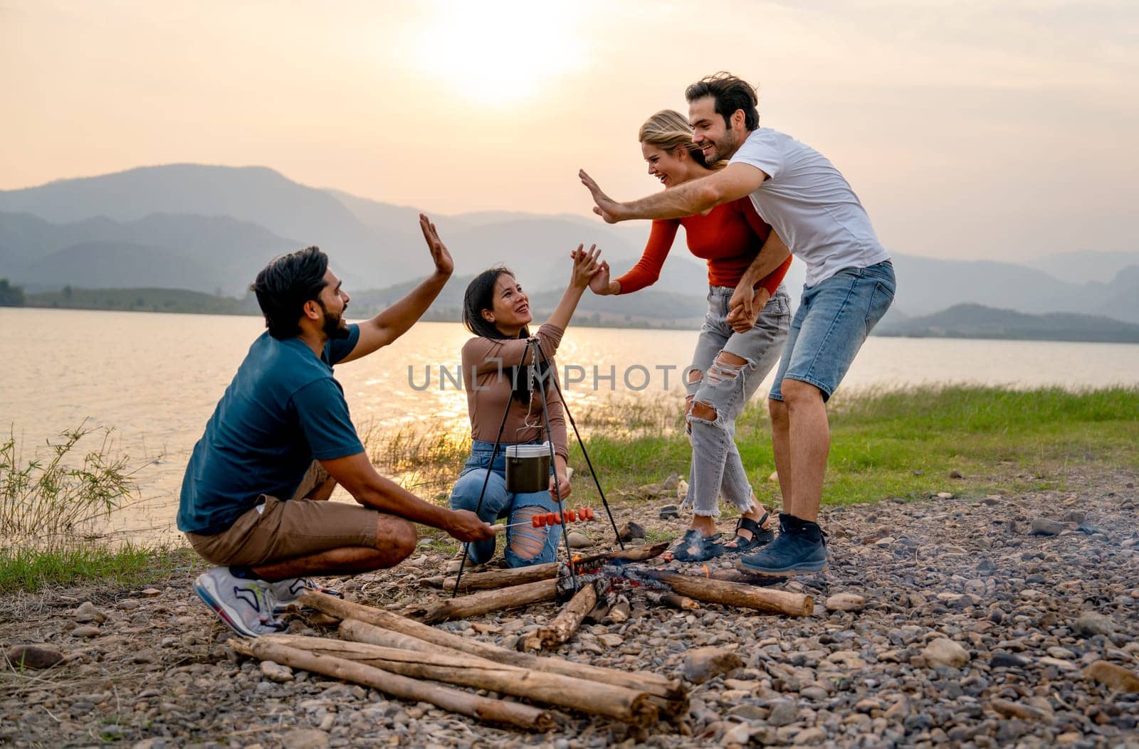 Caucasian man and woman walk and join and touch hands with Asian friends for party and camping near lake with sunset.