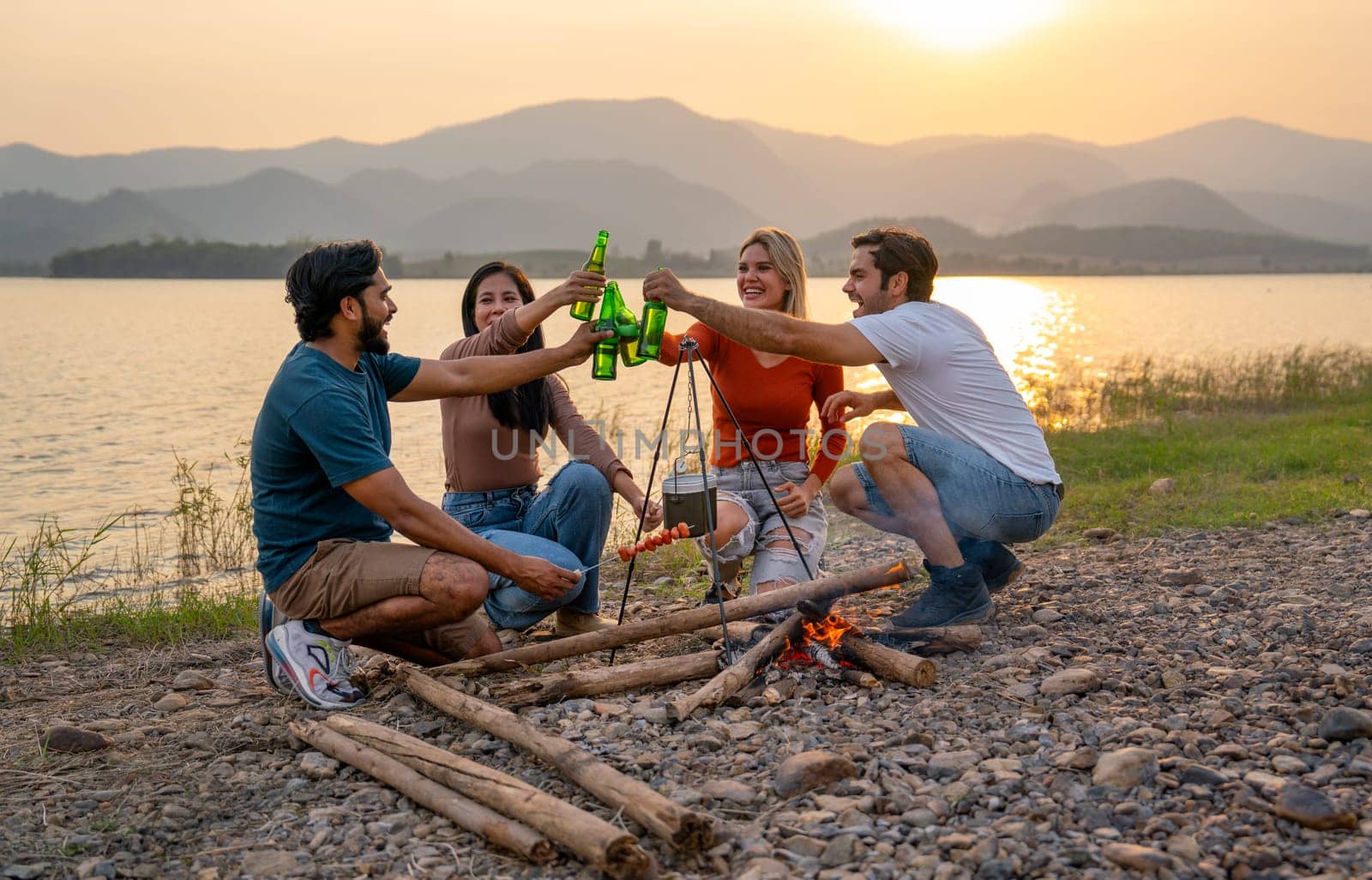 Group of men and woman friends with multi-ethnic enjoy in party and cheers action with surrounded by fire and they look happy to join together in public park near lake and sunset.