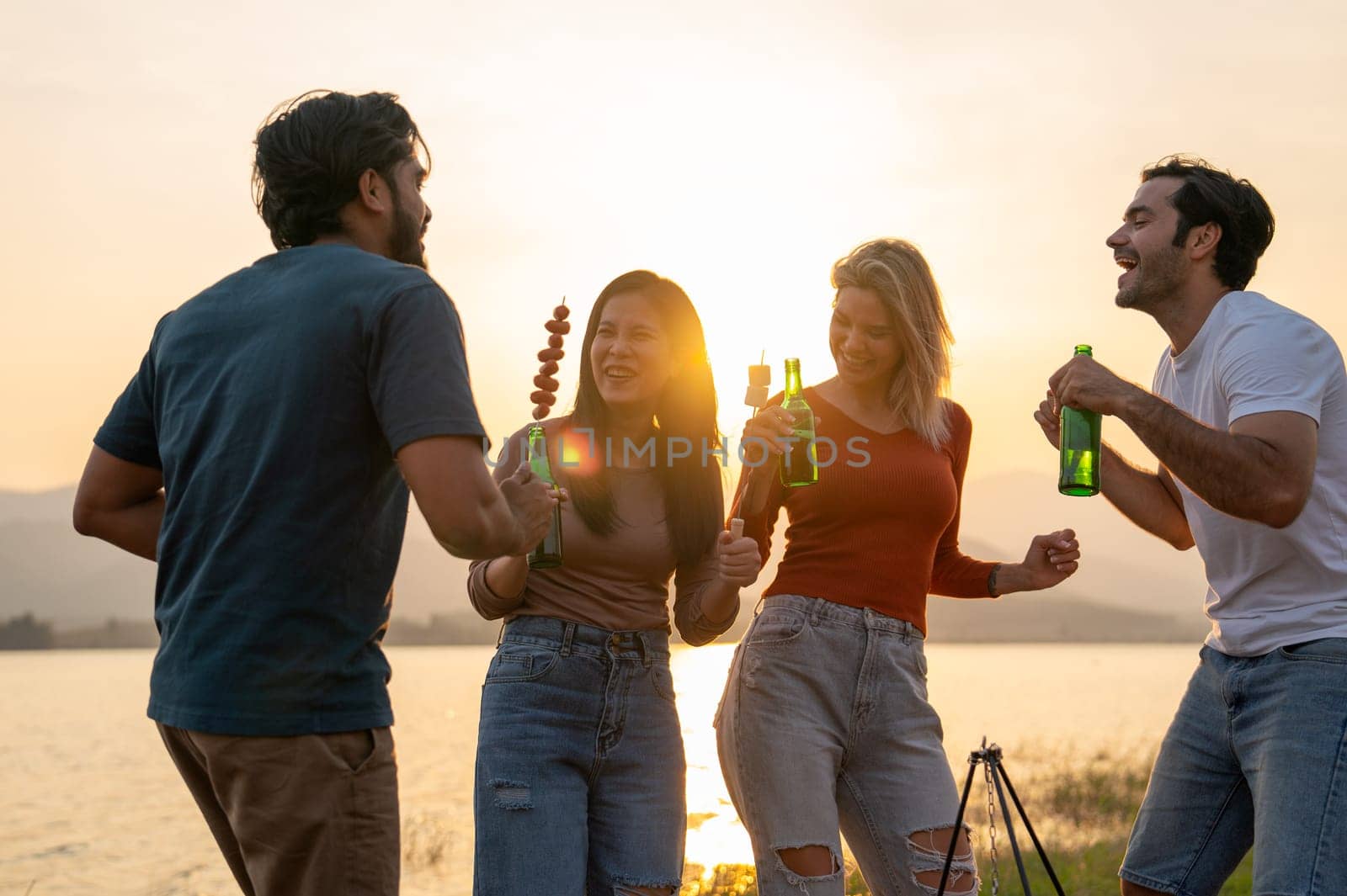 Group of friends with multi-ethnic enjoy with dancing and hold bottle of drinking in public park near the lake with sunset light.