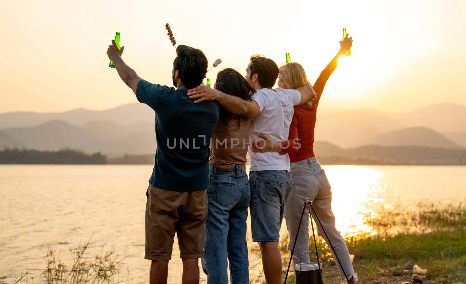 Back of group of friends with multi-ethnic hug together also hold bottle of drinking stand in front of sunset light near lak of public park with happiness. by nrradmin