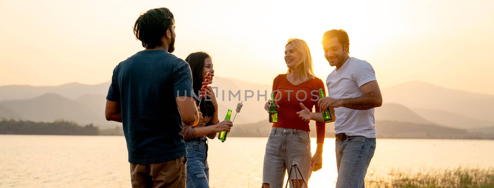 Group of men and women friends with multi-ethnic enjoy with talking and hold bottle of drinking in public park near the lake with sunset light.