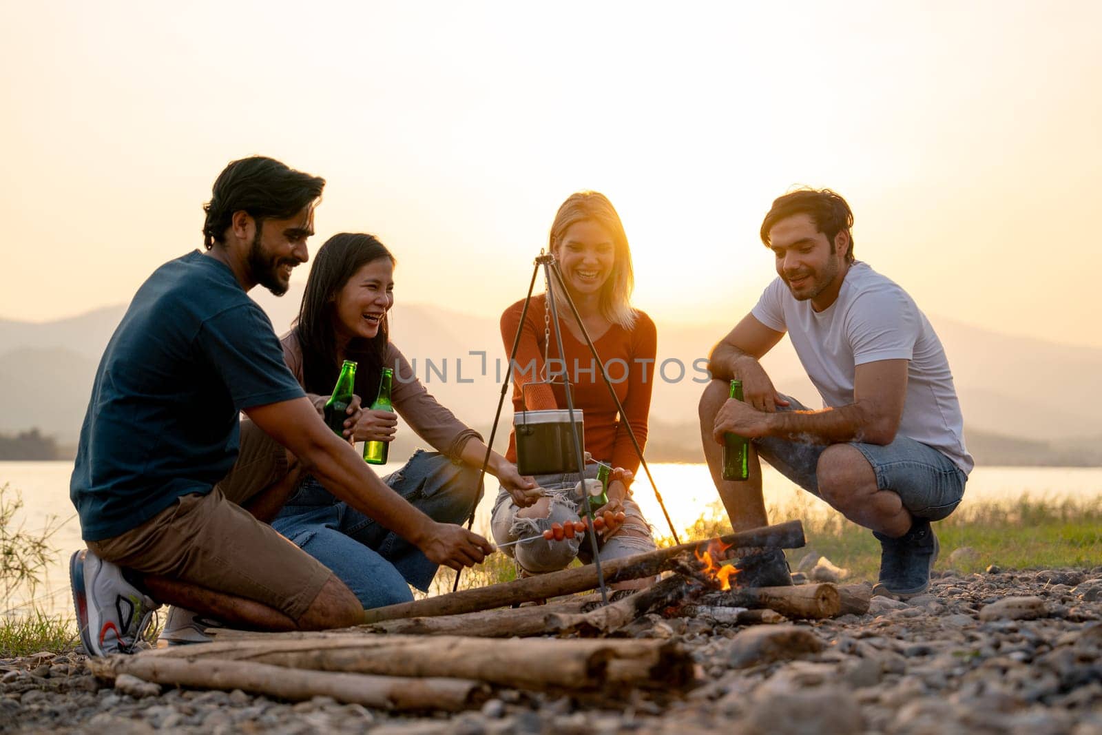 Group of men and woman friends with multi-ethnic enjoy to party and stay surrounded by fire and they look happy to join together in public park near lake and sunset. by nrradmin