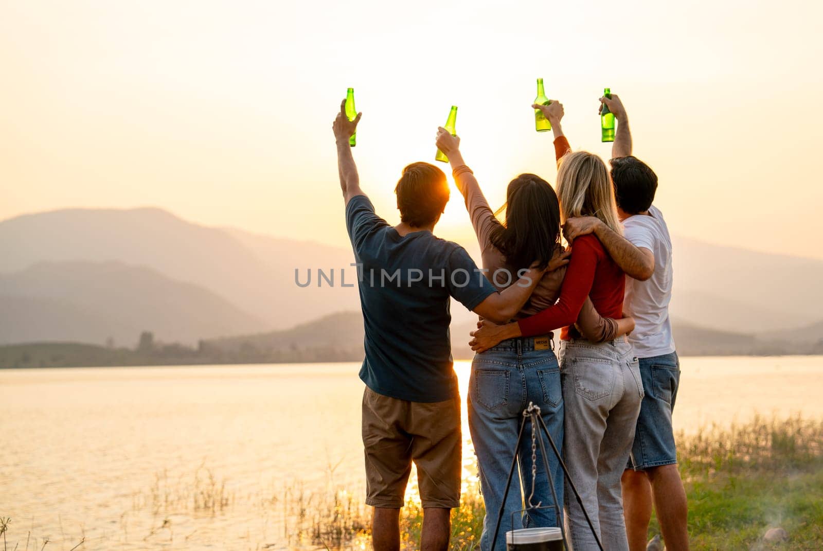 Wide shot of Back of group of friends with multi-ethnic hug together also hold bottle of drinking stand in front of sunset light near lake of public park.