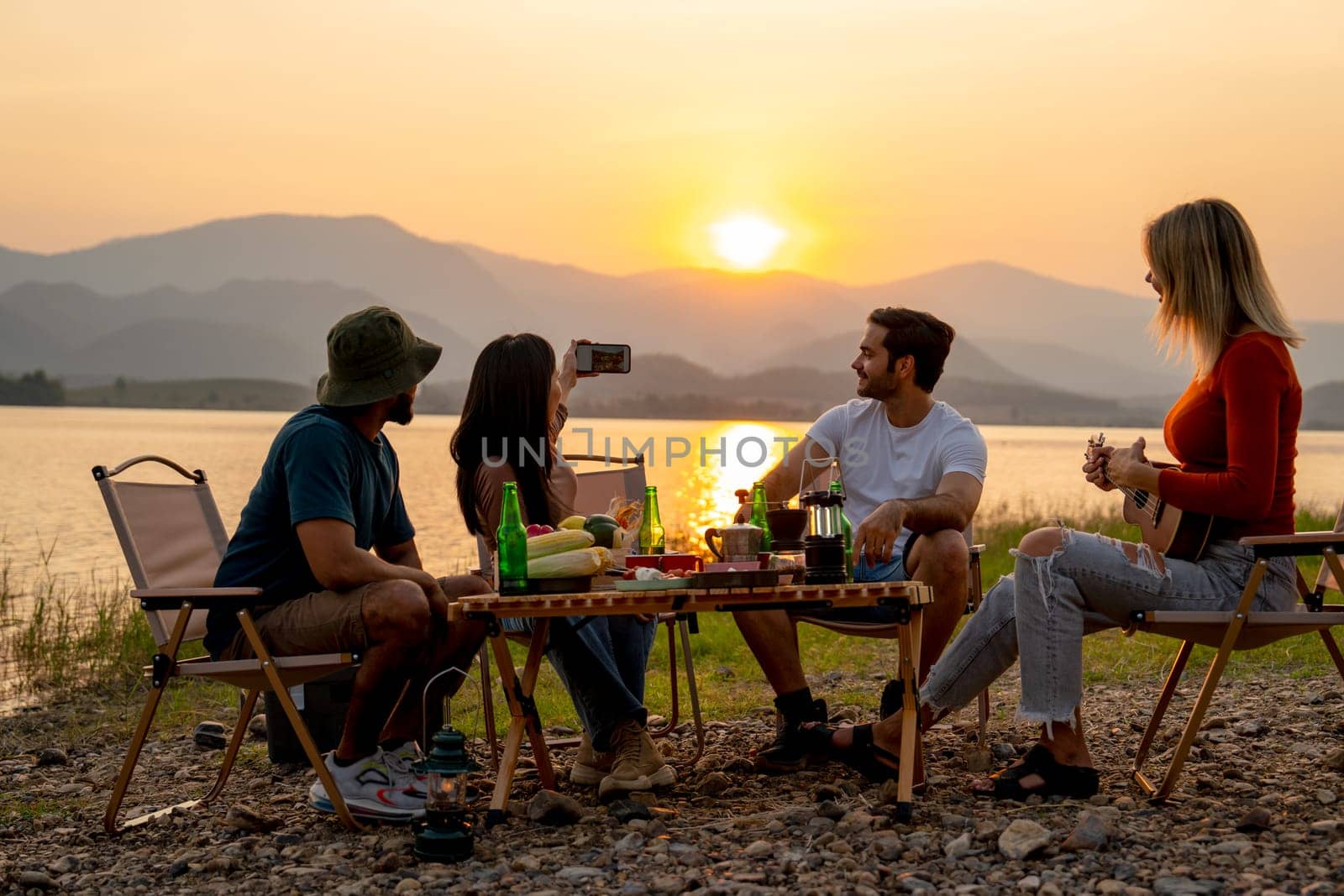 Beautiful Asian woman use mobile phone to selfie with her friends during camping and enjoy together near lake with sunset and they look happy for party outdoor. by nrradmin