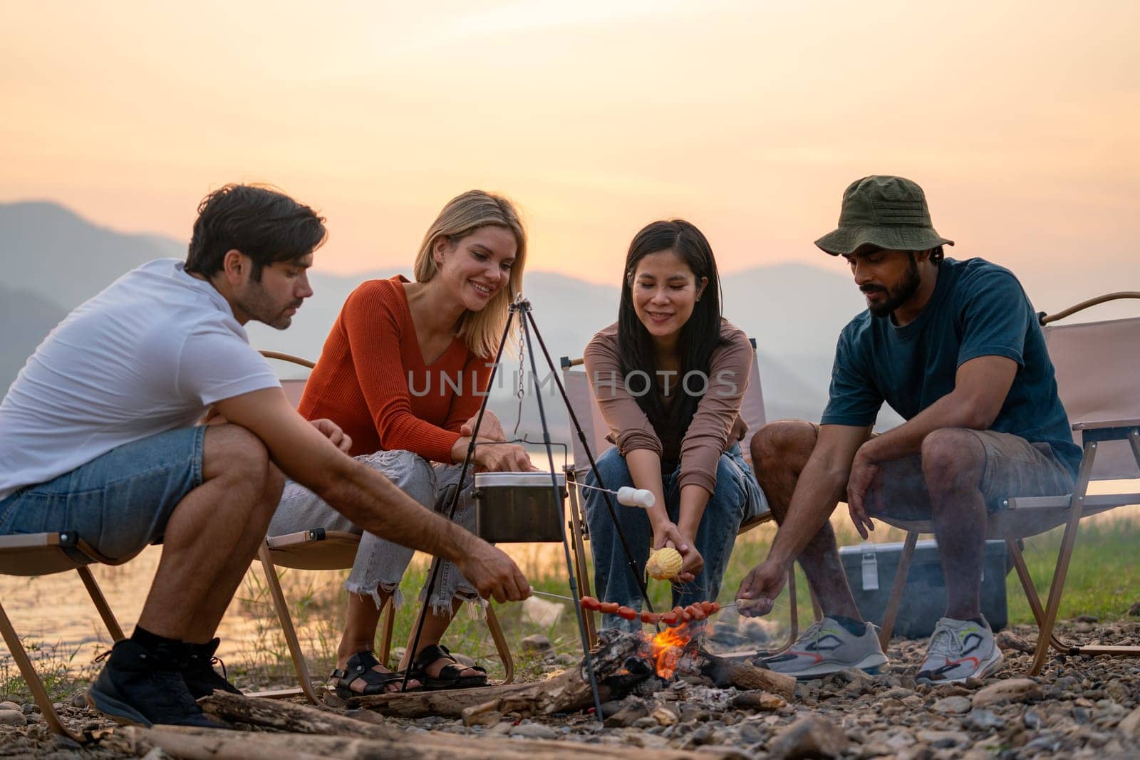 Close up group of men and woman friends with multi-ethnic enjoy to eat in party and sit surrounded by fire and they look happy to join together in public park near lake and sunset.