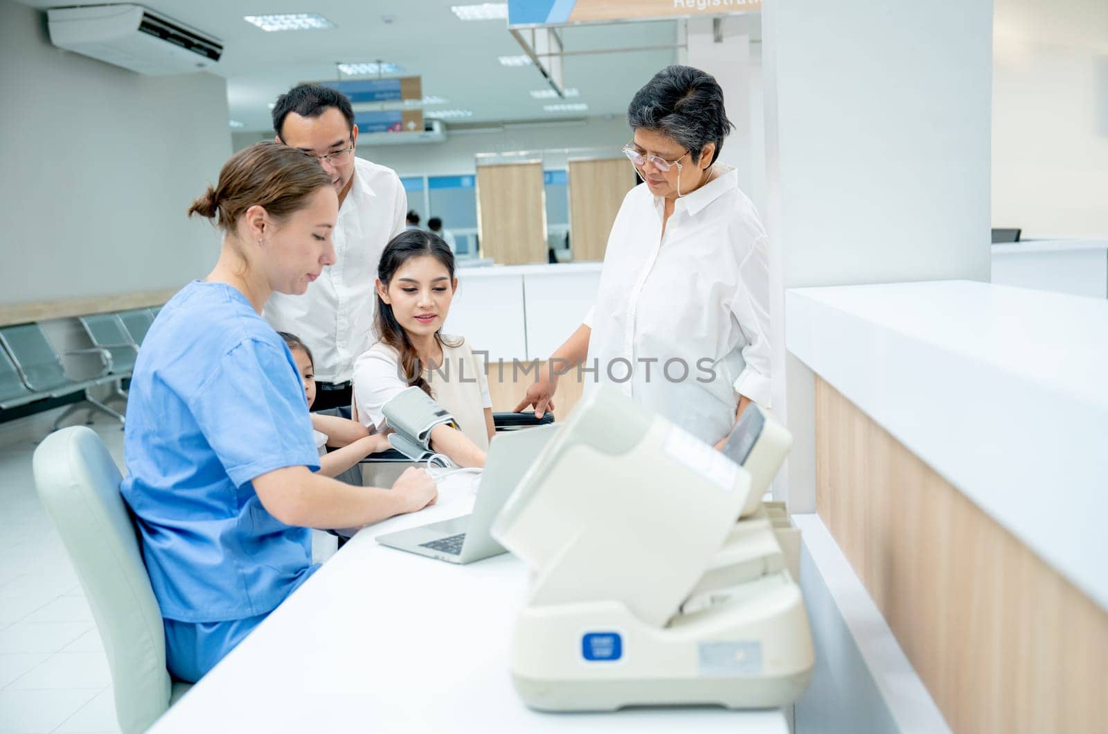 Caucasian nurse measure blood pressure of Asian woman and support by her family with husband, little girl and grandmother in area of hallway of hospital.
