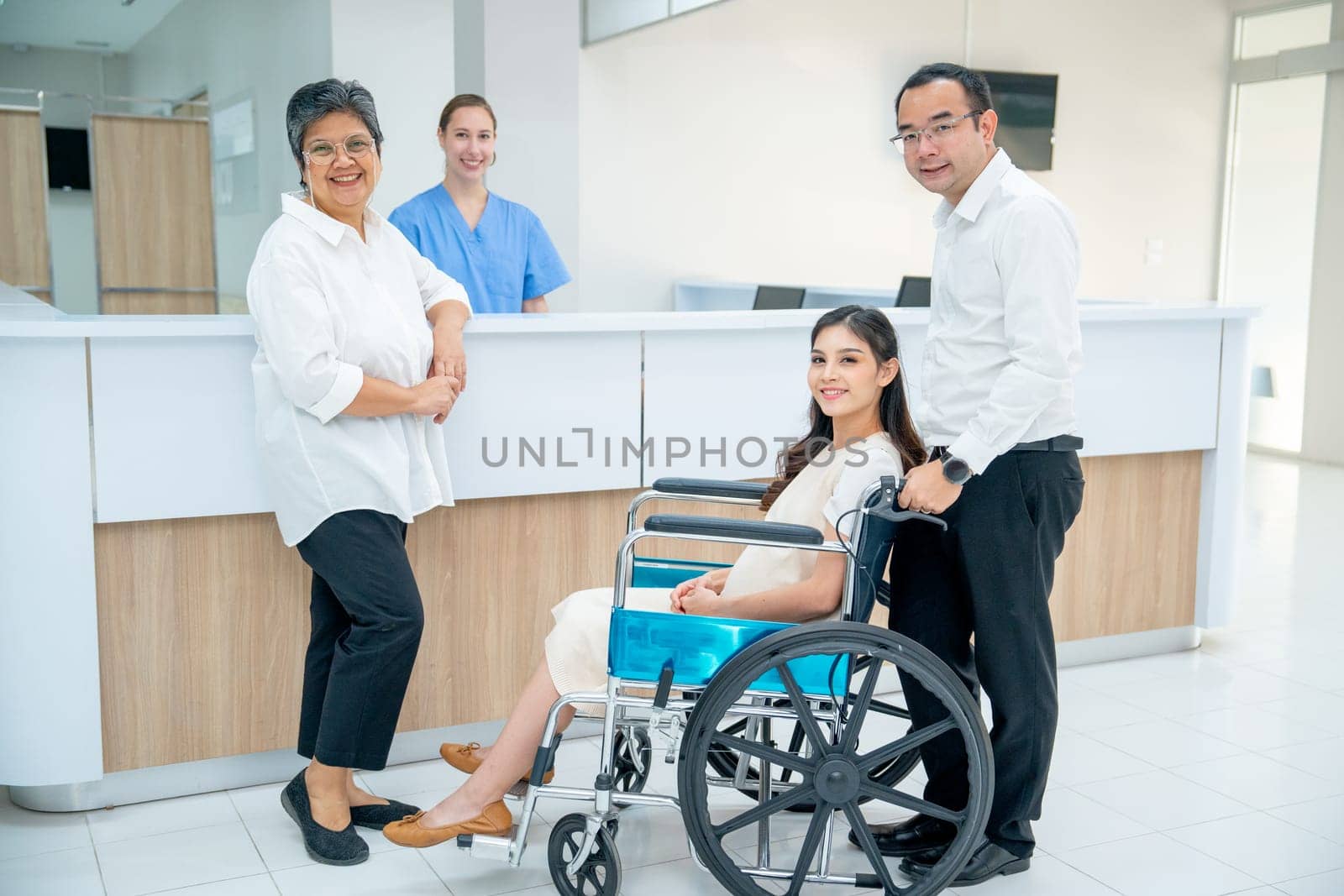 Asian family with senior woman, pregnancy woman sit on wheelchair and man take care the woman, they look at camera with nurse is smiling in the background in area of registration of hospital.