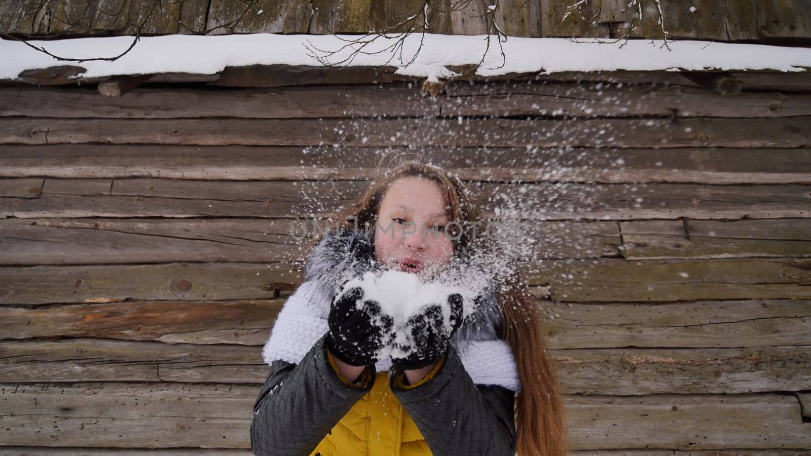 Young beautiful girl having fun blowing snow on the camera in winter clothes on the background of a wooden house in the village. Winter fun. by DovidPro