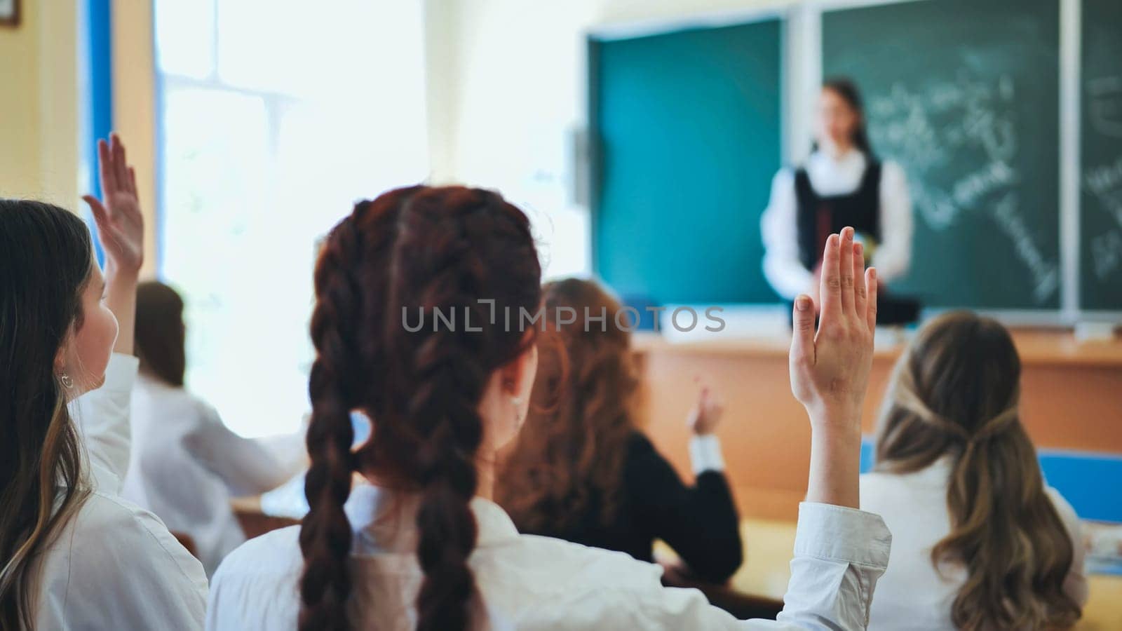 Intelligent group of young school children all raising their hands in the air to answer a question posed by the female teacher, view from behind by DovidPro