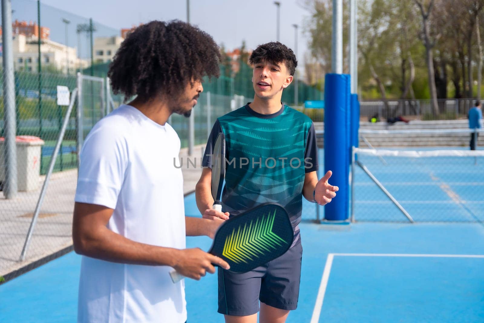 Caucasian young male instructor talking to a young african man playing pickleball in an outdoor court during sunny day