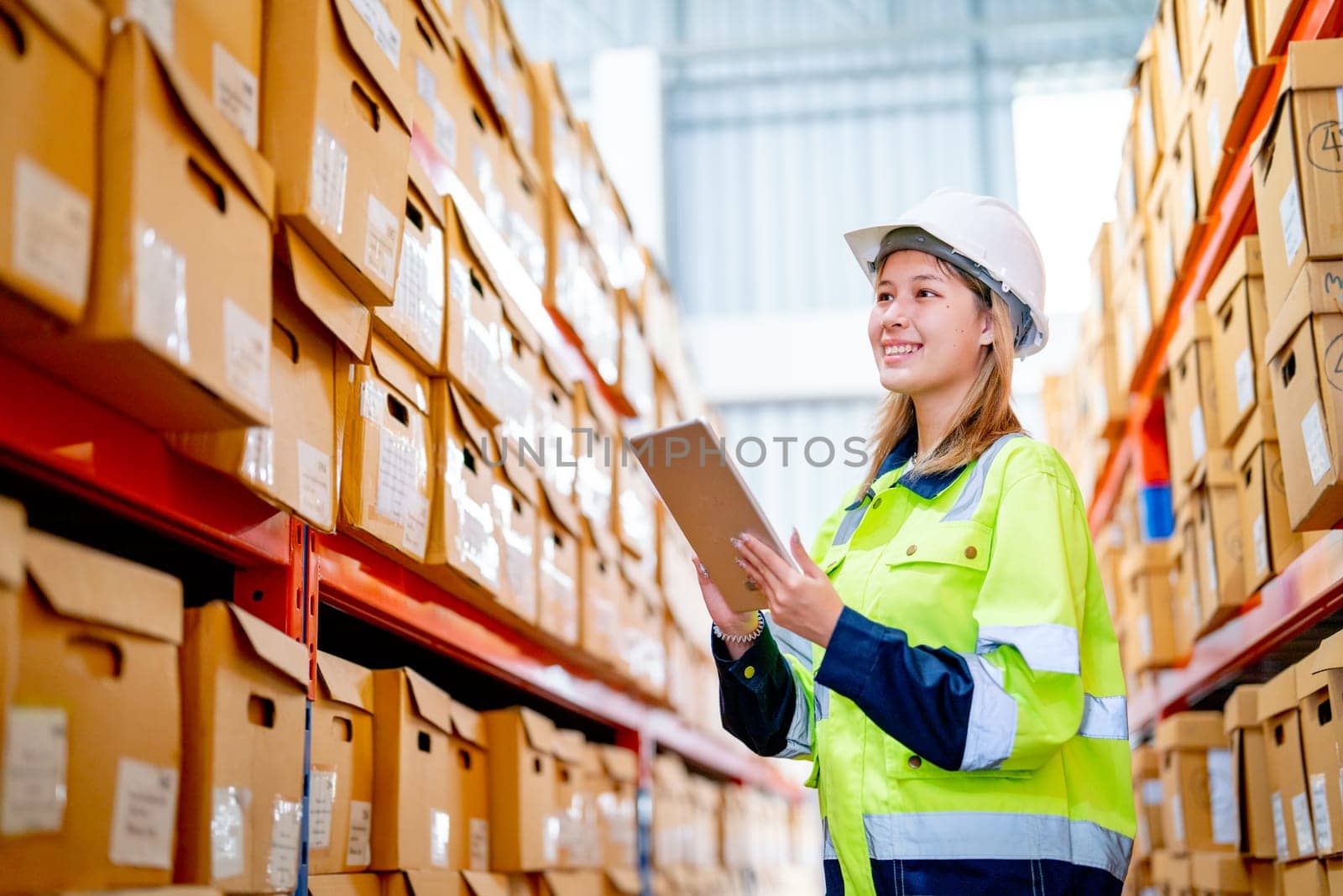 Pretty young warehouse worker woman hold tablet and look to product boxes on shelves during check stock in workplace area. by nrradmin