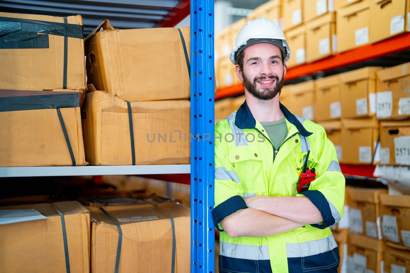 Smart warehouse worker man stand and lean to pole of product shelves also look at camera with smiling in workplace area. by nrradmin
