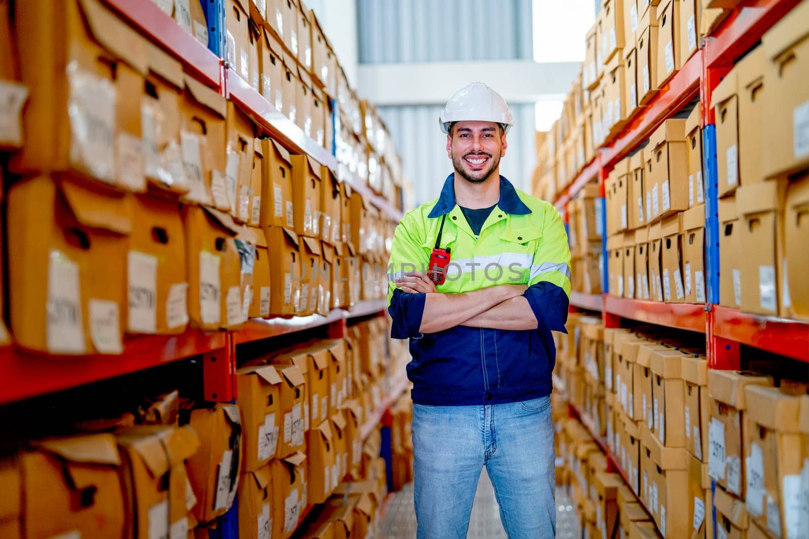 Professional warehouse worker man stand with arm-crossed and smiling to camera also stay between shelves of product boxes in workplace area.