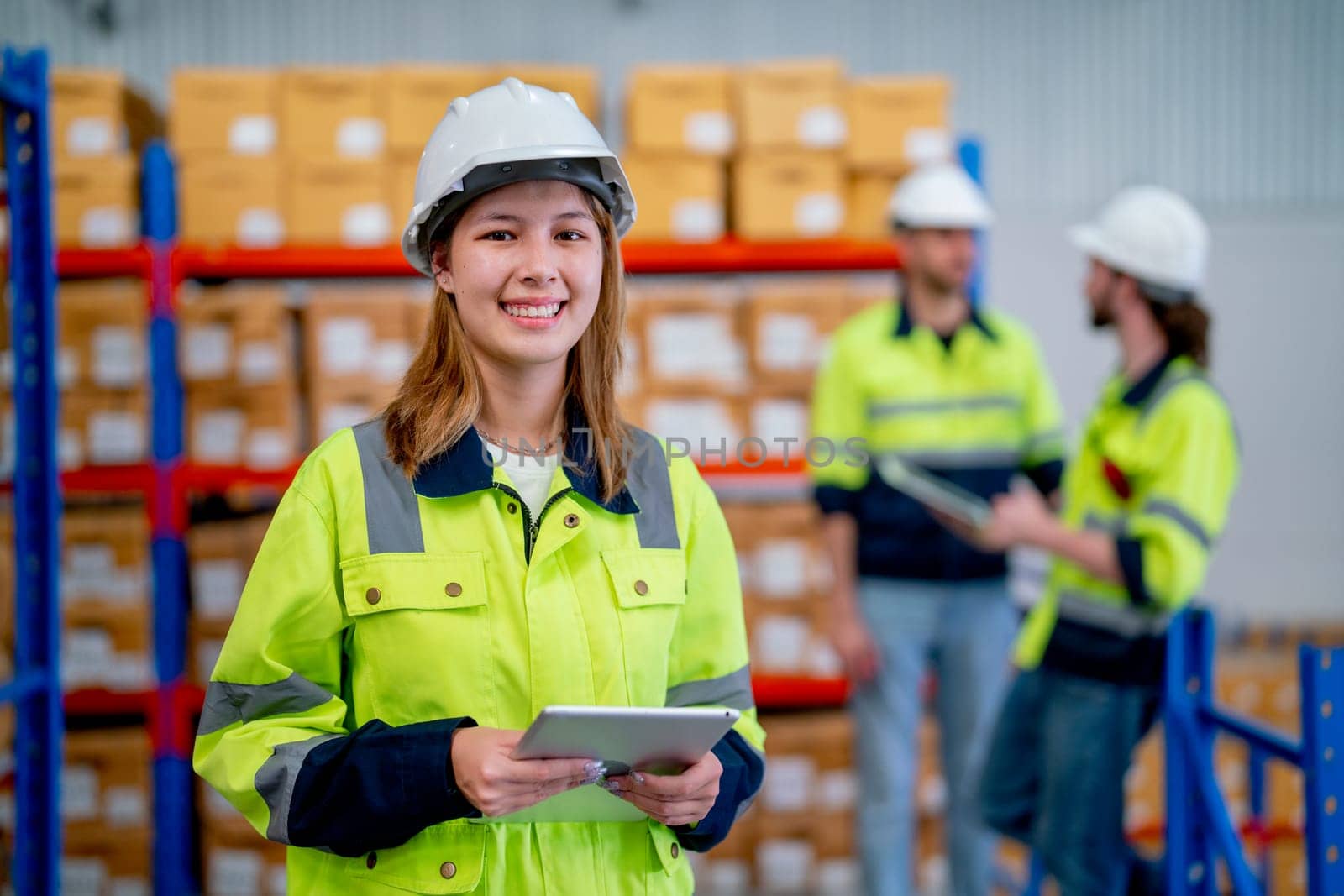 Warehouse worker young woman hold tablet and look at camera and smiling with her coworkers discuss in the background.