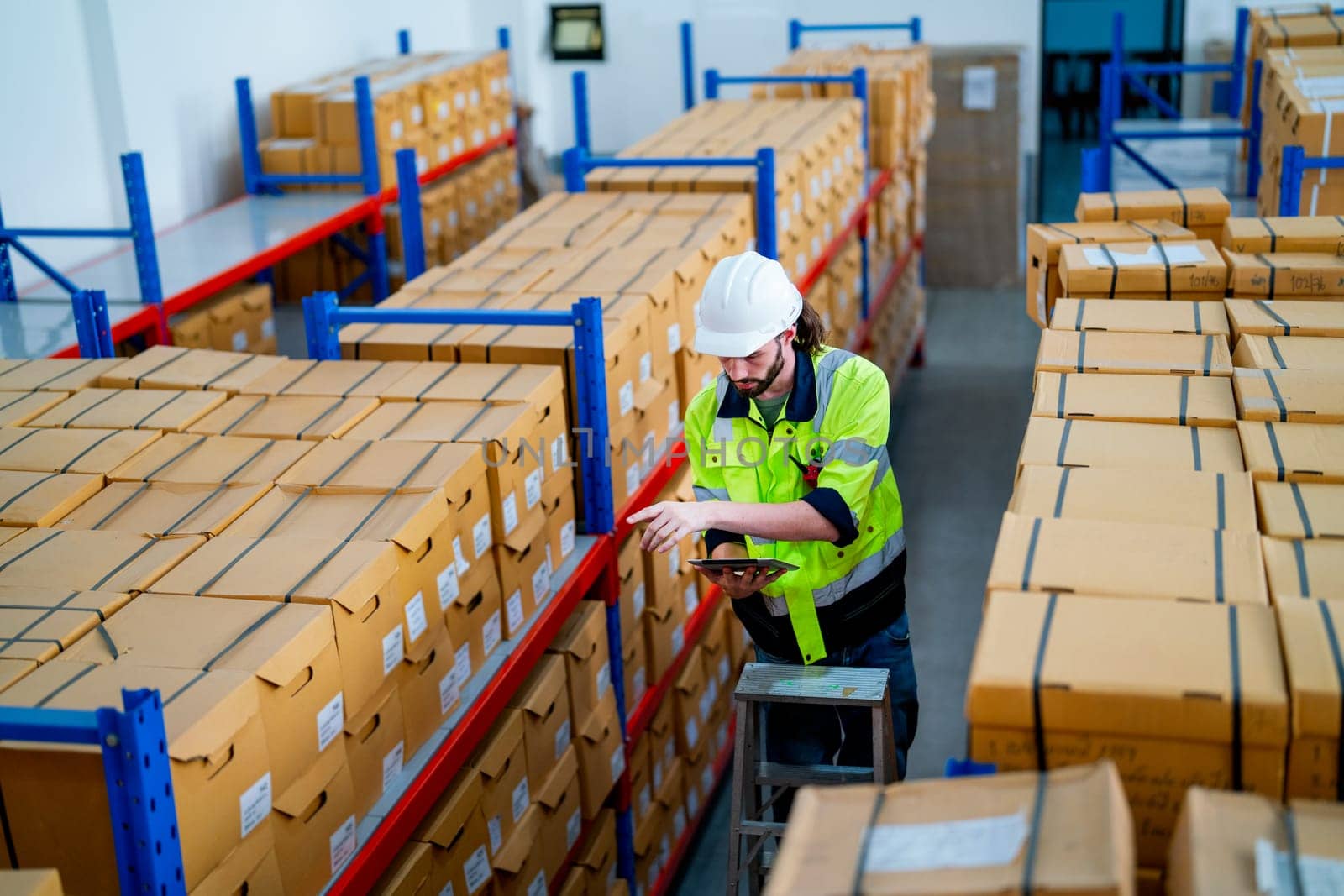 Upper view warehouse worker man stand on stair and hold tablet to check product on shelves in workplace.