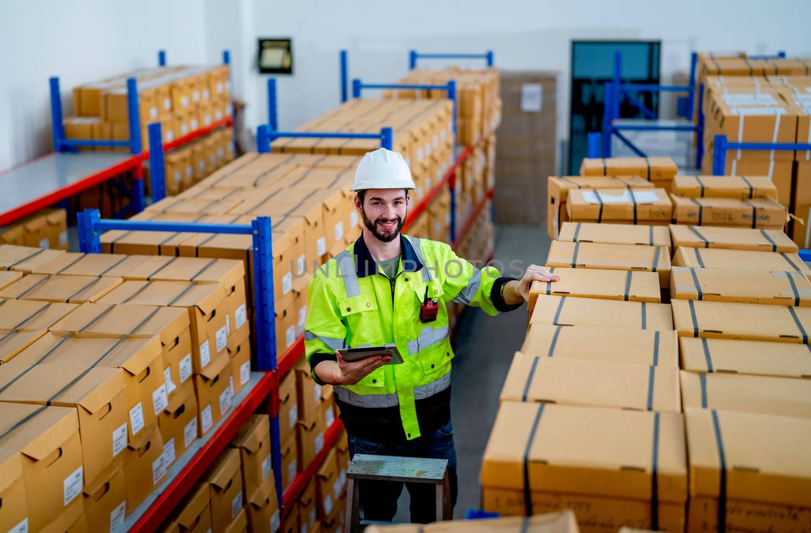 Warehouse worker man hold tablet and look at camera with smiling during stand on stair and check product on shelves in workplace.