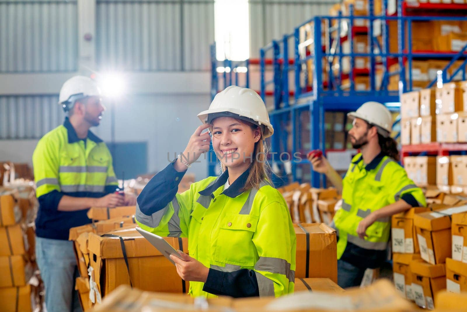 Pretty young warehouse worker woman touch her hair and look at camera with smiling also hold tablet with her co-worker discuss in the background. by nrradmin