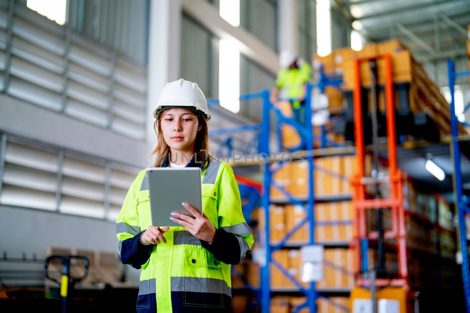 Pretty young warehouse worker woman hold tablet and look at tablet and her co-worker work on the background during carry product out from cargo truck in workplace.