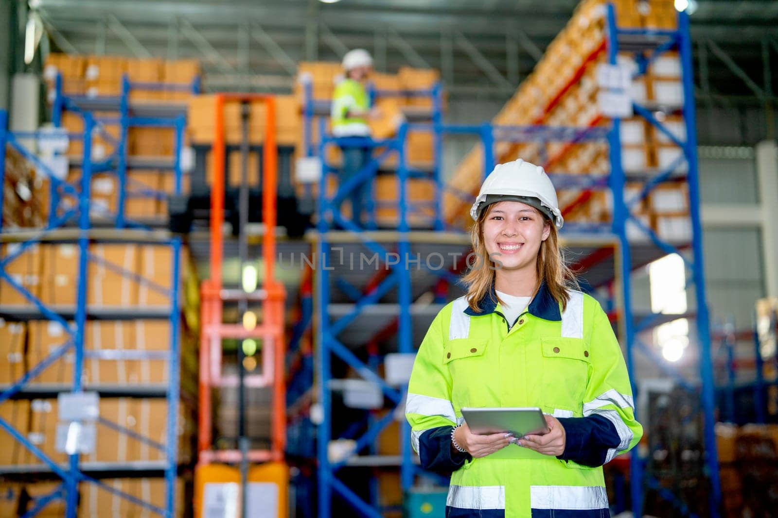 Portrait of pretty young warehouse worker woman hold tablet and look at camera with smiling and her co-worker work on the background during carry product out from cargo truck in workplace.