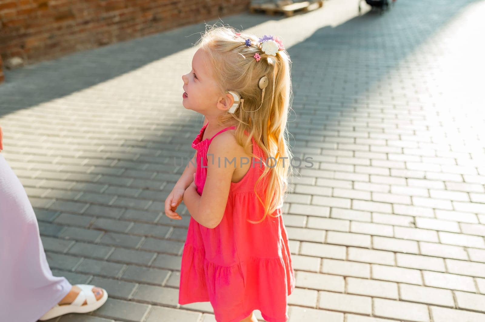 Child girl with hearing aid cochlear implant in pink dress having fun on summer street. Aid for the treatment of deafness and hearing loss in humans. Copy space by Satura86