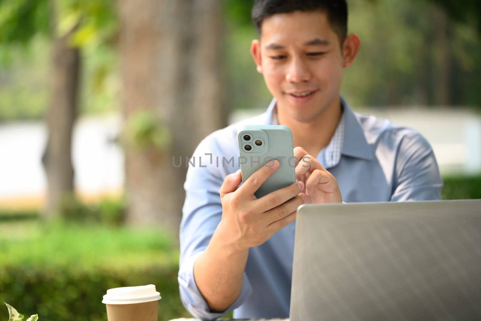 Smiling young businessman texting message on mobile phone sitting outside at cafe table.