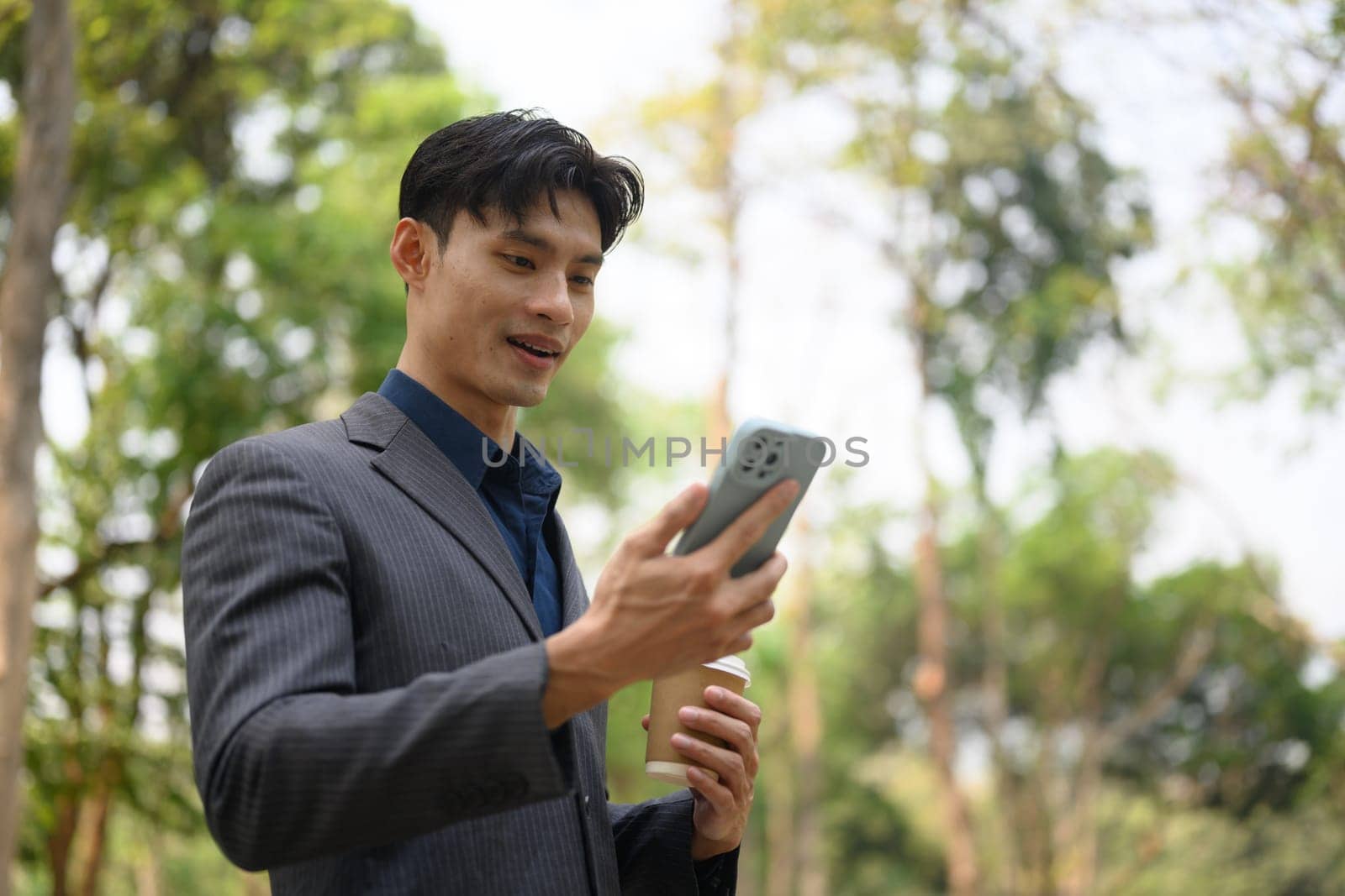 Handsome businessman with paper cup of coffee walking in city park and using smartphone.