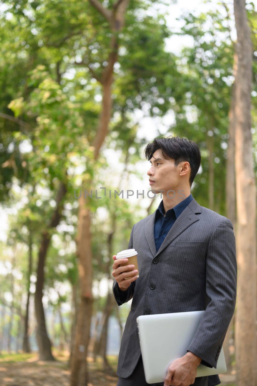 Portrait of handsome businessman holding paper cup of coffee standing in the public park.