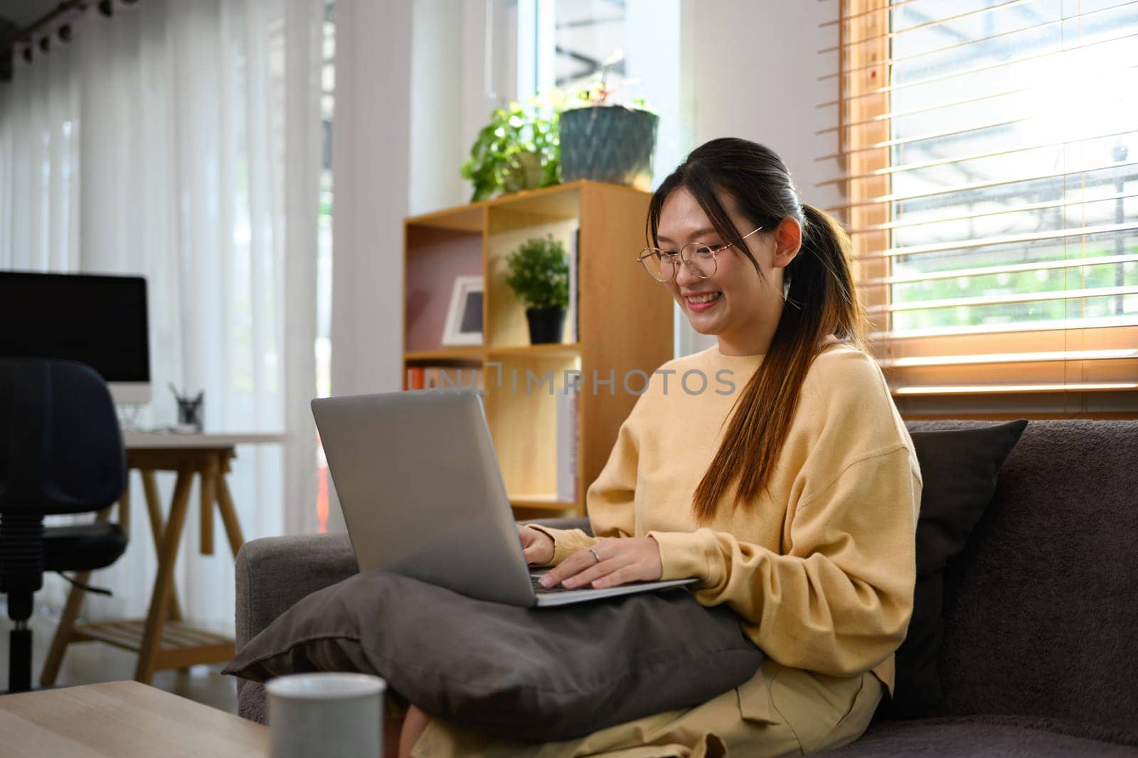 Young woman in casual clothes using laptop sitting on sofa at cozy home.