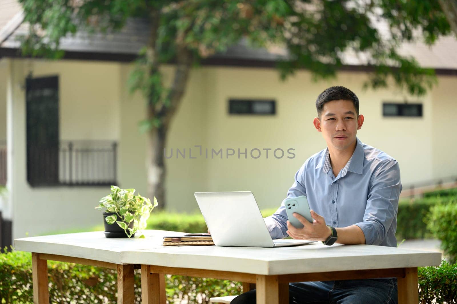 Handsome young businessman using laptop outdoors, holding smartphone and looking away.