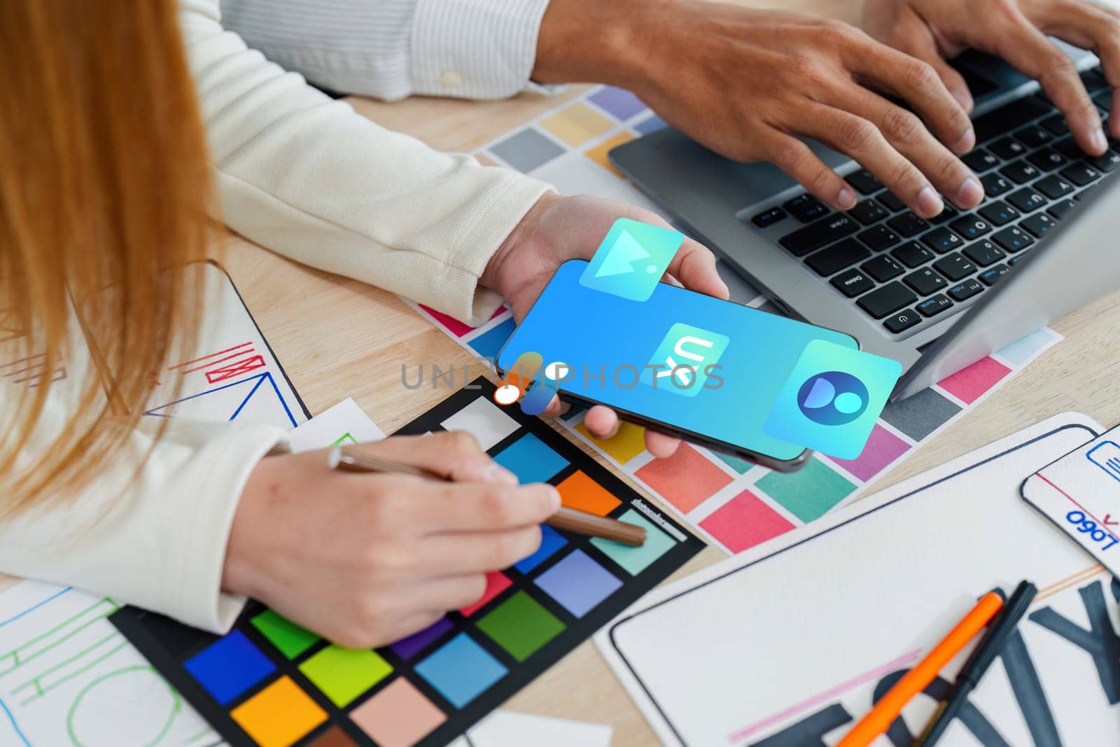 Two people are working on a project together, one of them is holding a cell phone and the other is holding a pen. They are sitting at a table with a laptop and a stack of papers