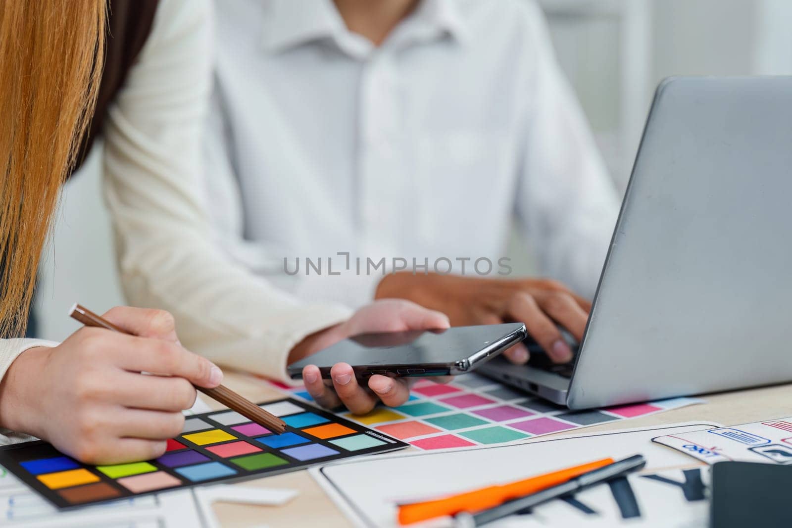 Two people are working on a project together, one of them is holding a cell phone and the other is holding a pen. They are sitting at a table with a laptop and a stack of papers